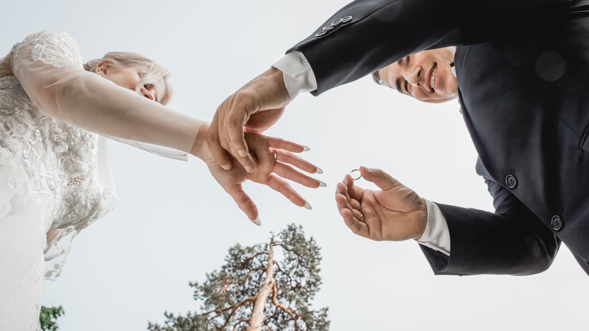 Couple exchanging wedding rings outdoors under a clear sky.