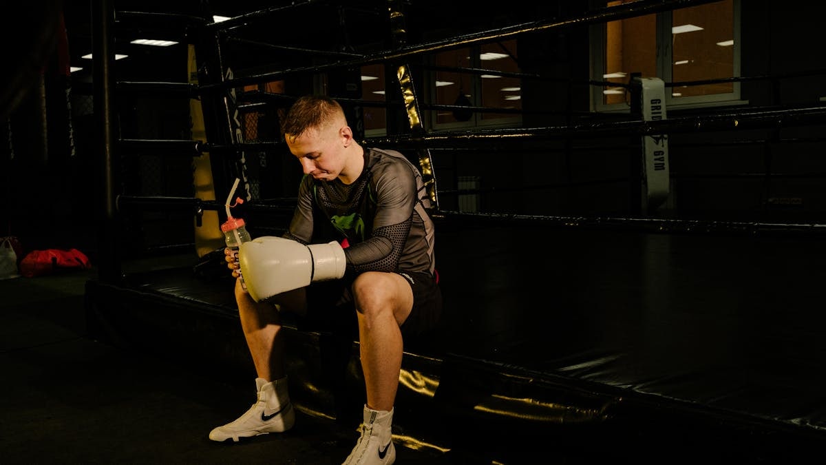 Young male boxer sitting in the ring holding gloves and a water bottle, taking a break.